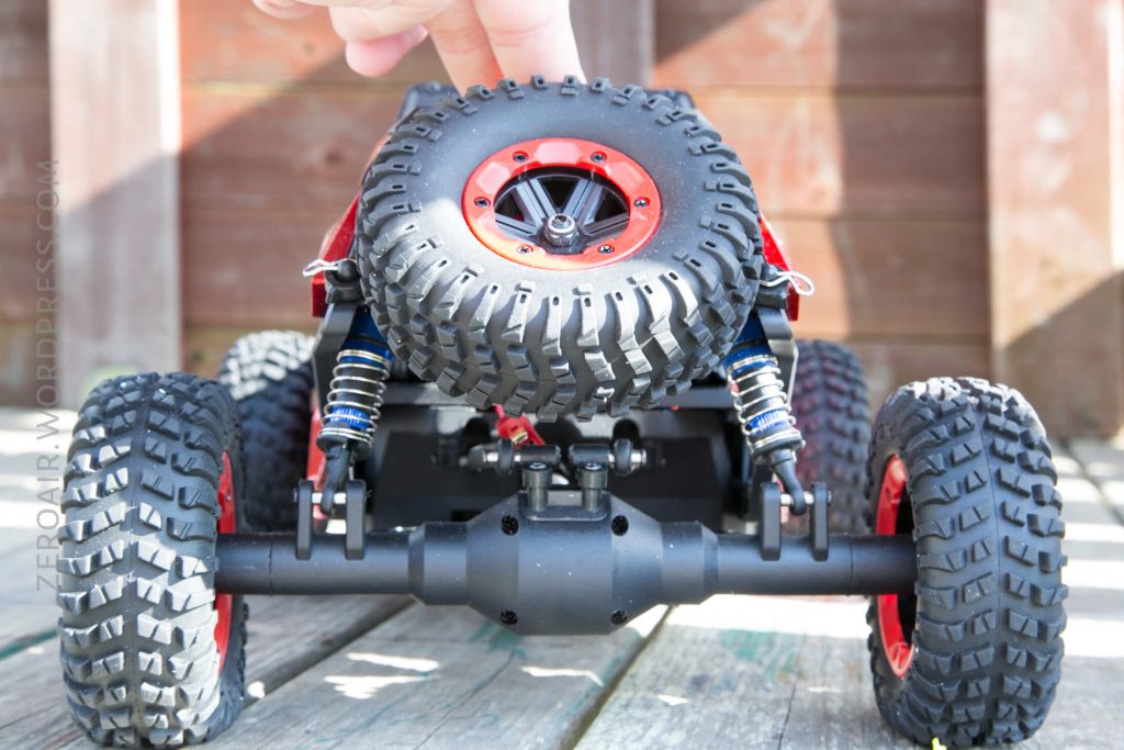 A close-up view of the rear side of a remote control off-road vehicle with large, rugged tires and red accents. A hand is touching the spare tire mounted on the back. The vehicle is on a wooden surface.