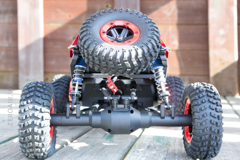 Close-up rear view of a remote-controlled off-road vehicle with large, knobby tires, visible suspension springs, and a mounted spare tire. The car is positioned on wooden planks with a wooden fence in the background.