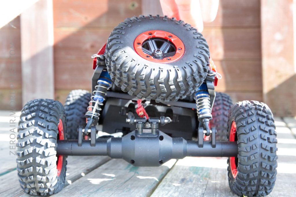 A close-up view of the underside of a remote control car with large, knobby tires and visible suspension springs. A hand is holding the car, and it is placed on wooden boards.