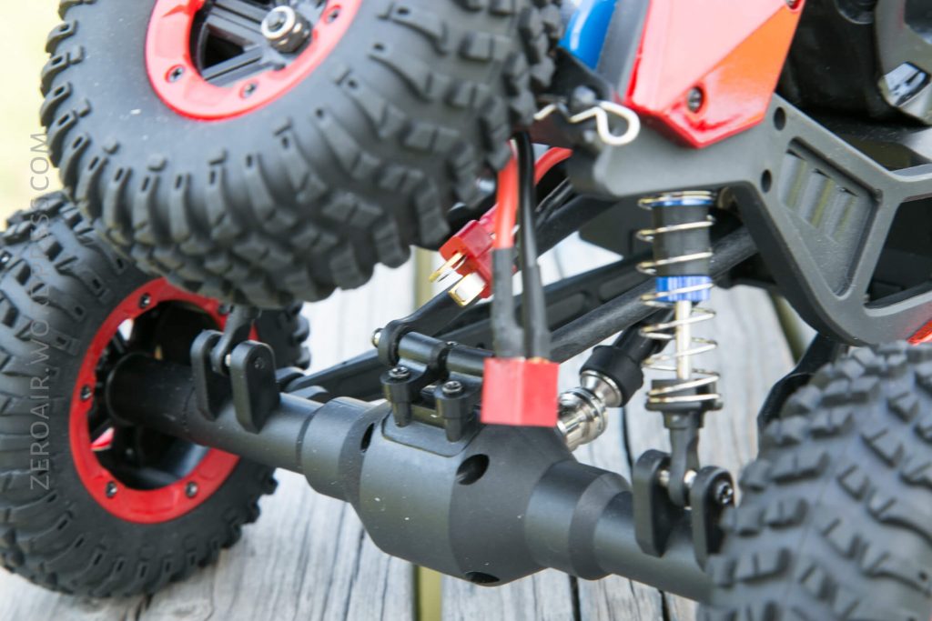 Close-up of the suspension and axle components of a red and black remote control off-road vehicle on a wooden surface. The image shows detailed parts including the shock absorber, tires, and chassis.