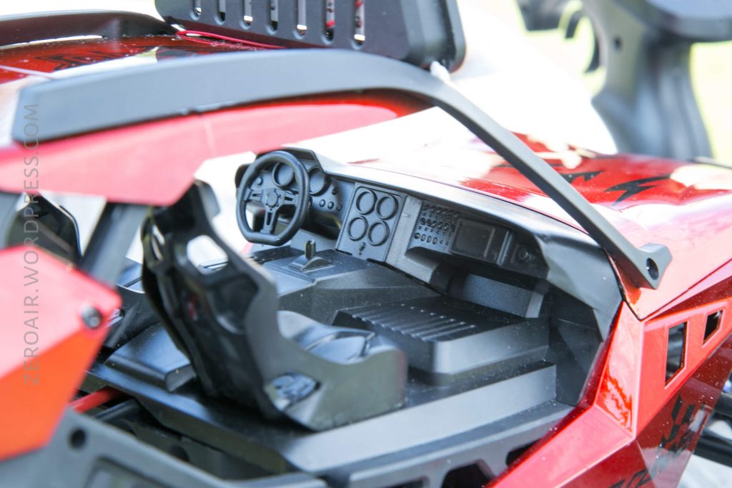 Close-up view of the interior of a red toy or model car, showing a detailed dashboard, steering wheel, and seat. The background is out of focus, and part of the exterior frame is visible.