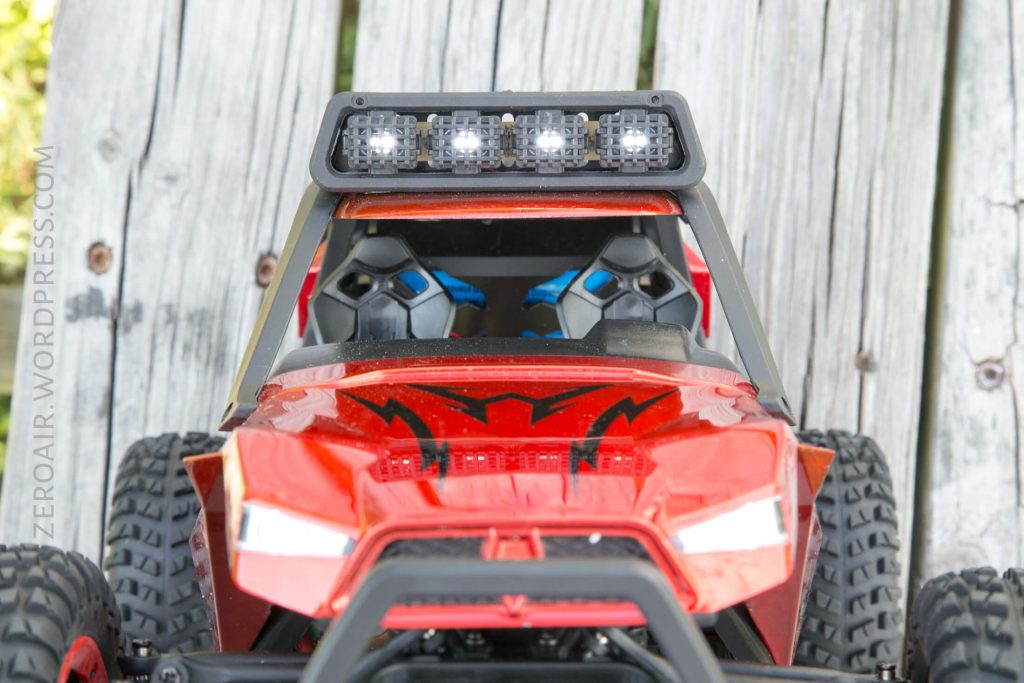 A close-up of a red remote-control car with a black grill, LED light bar on the roof, and two empty seats visible through the windshield, photographed on a weathered wooden surface.