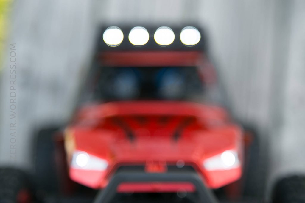 A blurred close-up image of a red vehicle, possibly a toy car or remote-controlled car, with four bright lights on top and a gray background. The details of the vehicle are not clearly visible.