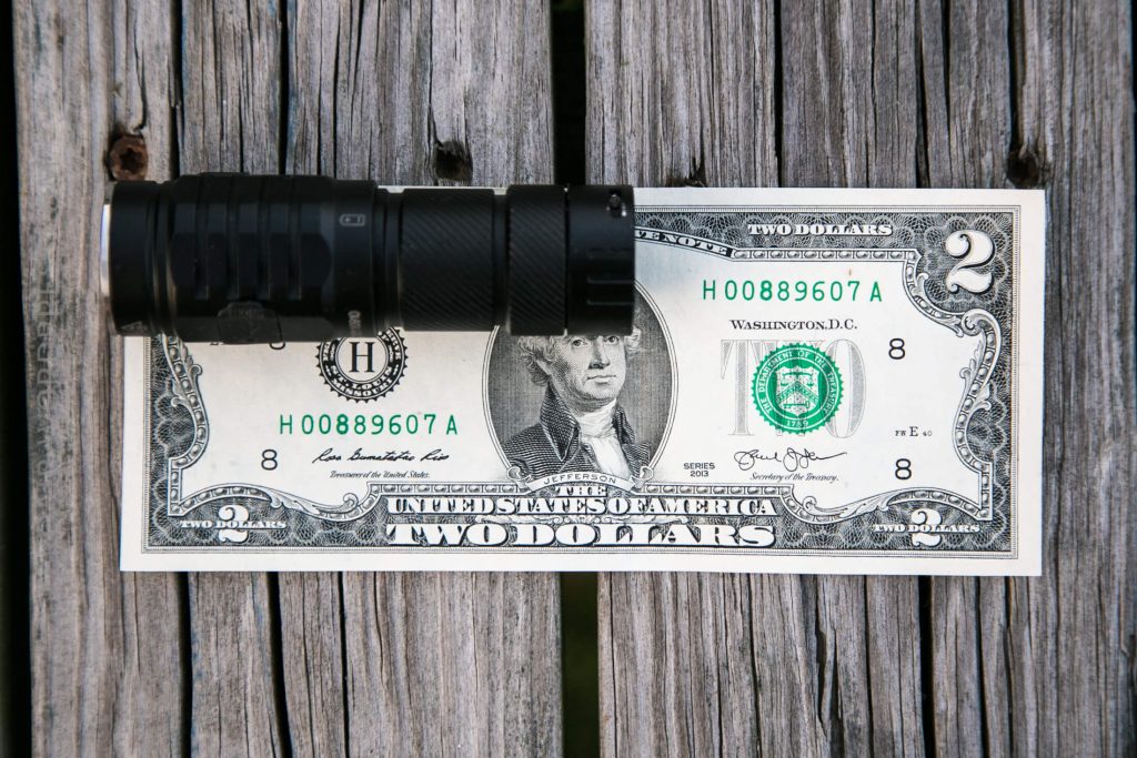 A black flashlight is placed horizontally on top of a two-dollar bill lying on a wooden surface.