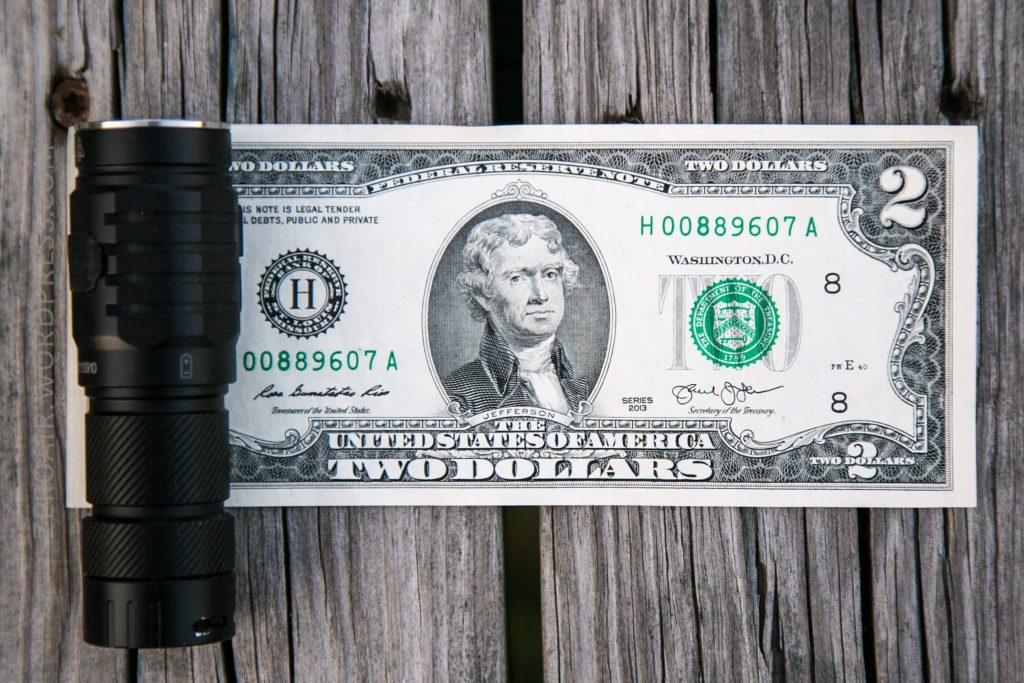 A black flashlight is placed next to a United States two-dollar bill on a wooden surface. The bill features a portrait of Thomas Jefferson and is positioned horizontally.