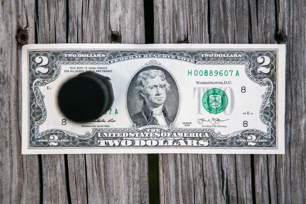 A two-dollar bill lies on a wooden surface with a black circular object placed over the left side, partially covering Thomas Jefferson’s portrait. The bill’s serial number is visible.