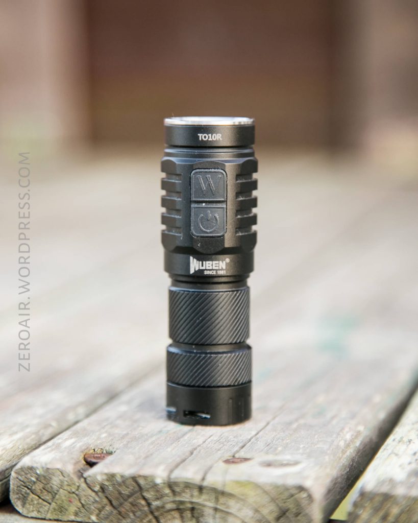 A black Wuben TO10R flashlight standing upright on a weathered wooden surface, with a blurred outdoor background and the watermark “ZEROAIR.WORDPRESS.COM” along the left side.