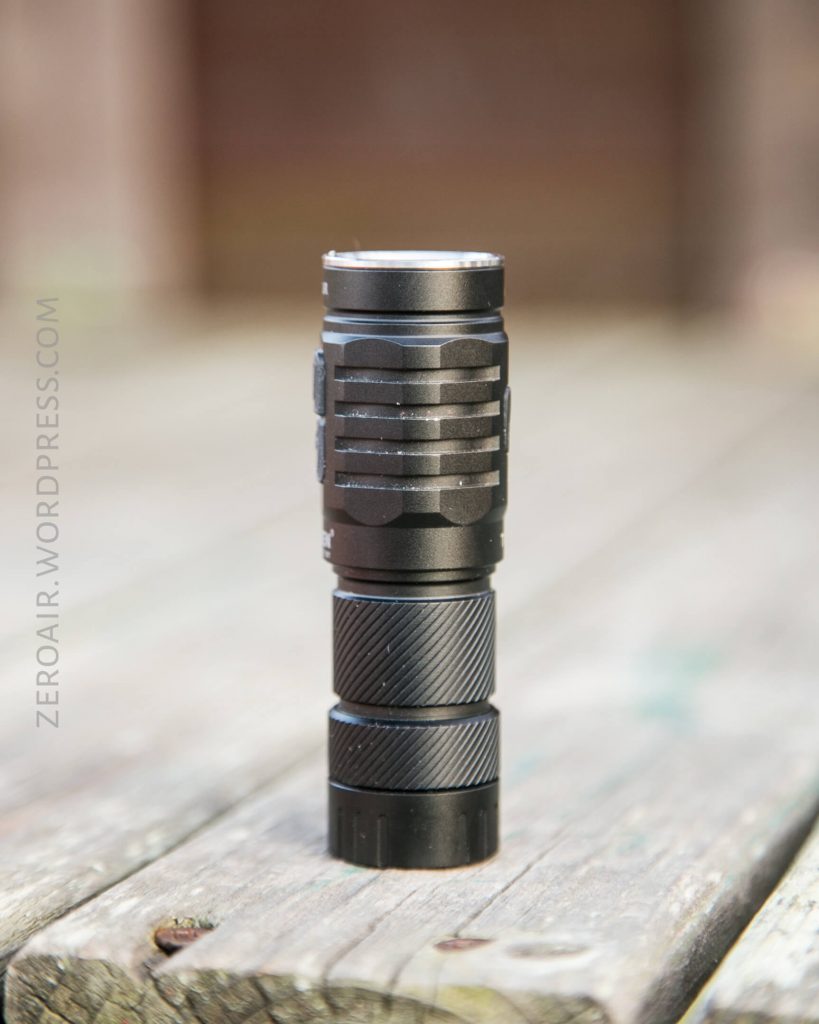 A small black flashlight stands upright on a weathered wooden surface. The flashlight has a textured grip and ridged side panels. The background is blurred. The text ZEROAIR.WORDPRESS.COM appears along the left side.