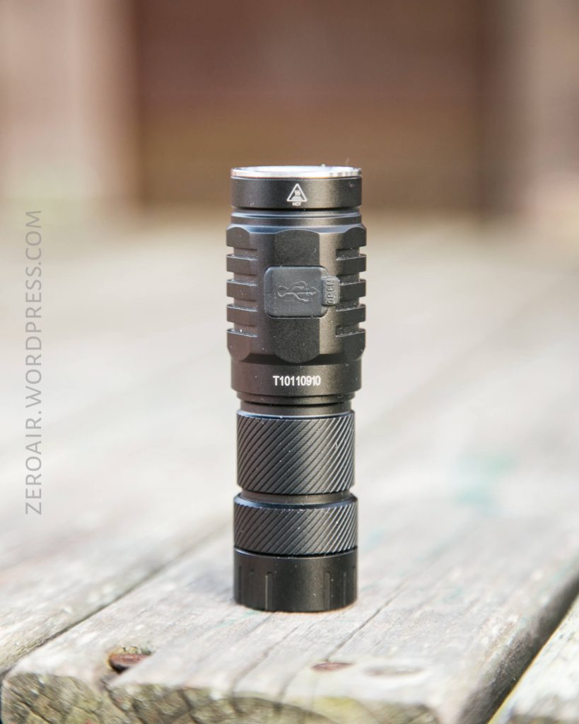 A black flashlight with textured grip and USB charging port is standing upright on a weathered wooden surface. The flashlight model number “T1010910” is visible on its body.