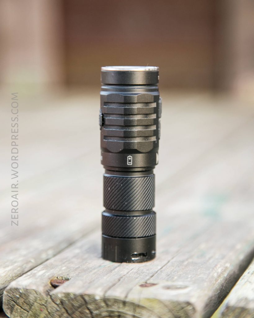 A black compact flashlight standing upright on a weathered wooden surface with a blurred background. The flashlight features a textured grip and visible control button.