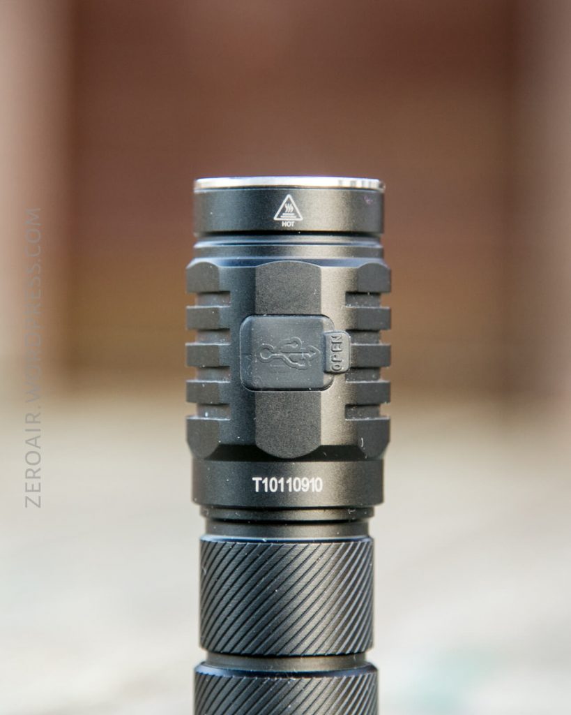 A close-up view of a black tactical flashlight standing vertically on a blurred surface, showing the model number T10110910 and ridged texture on the grip.