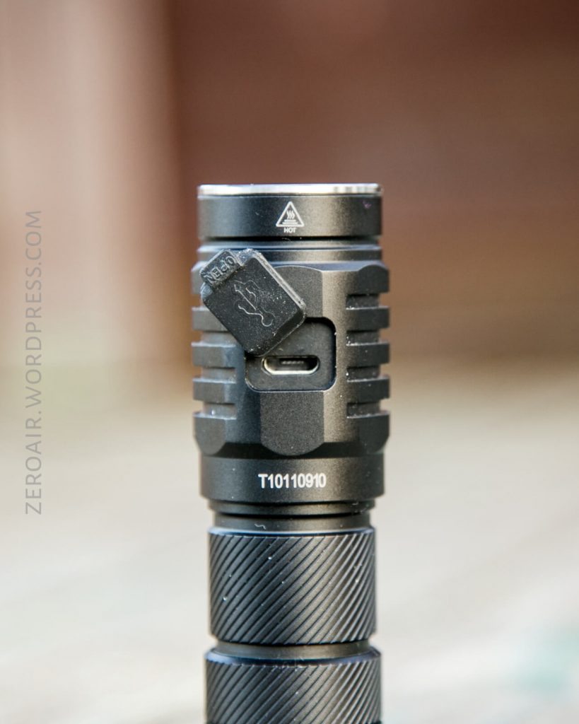 A close-up of a black flashlight showing a USB-C charging port covered by a rubber flap. The flashlight has a textured grip and ridged design. The background is blurred.