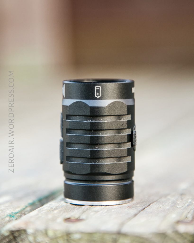 A small, cylindrical black flashlight with ridged grip is standing upright on a weathered wooden surface. The background is blurred, and the text ZEROAIR.WORDPRESS.COM is visible along the left side of the image.