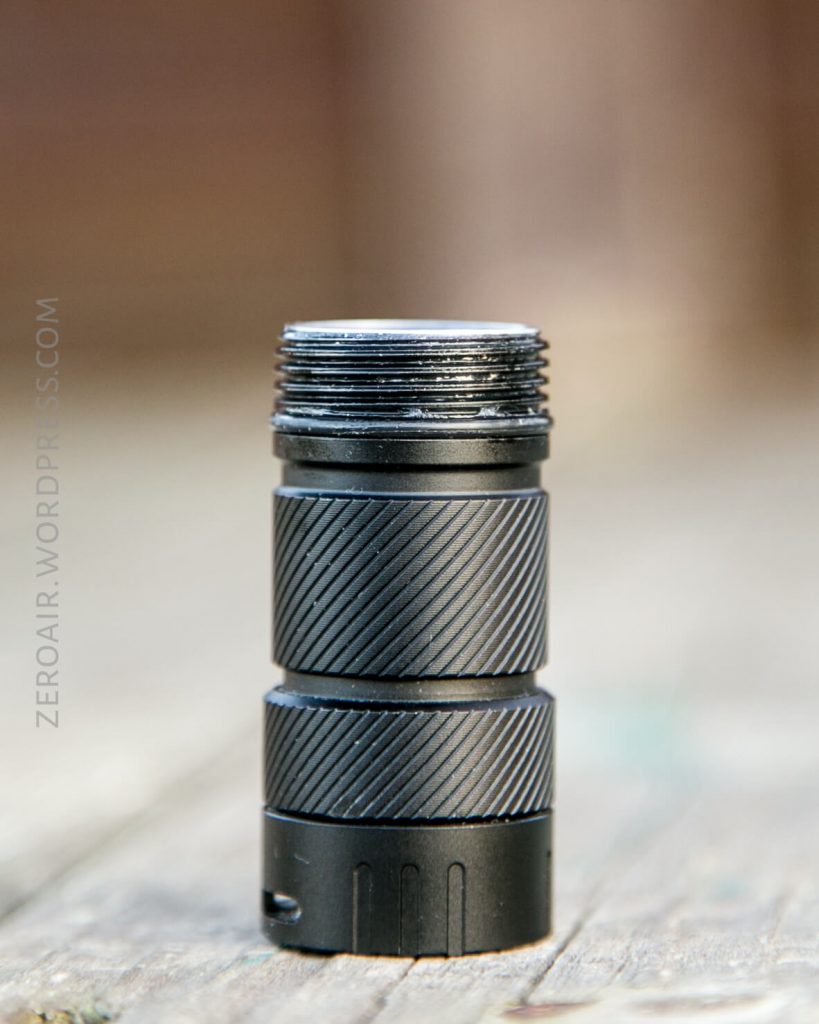 A close-up of a black metal flashlight body with visible knurled grip and open, threaded top, standing vertically on a wooden surface. The background is out of focus.