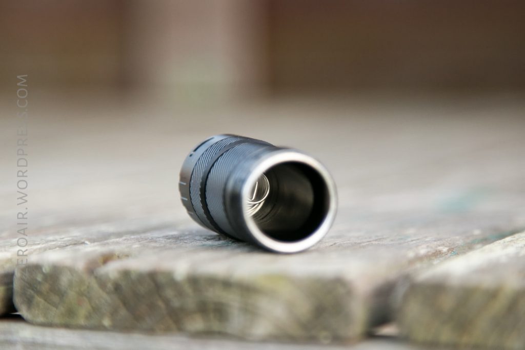 A close-up view of a cylindrical metal object, likely a flashlight body, lying on its side on a wooden surface. The inner spring is visible, and the background is blurred.