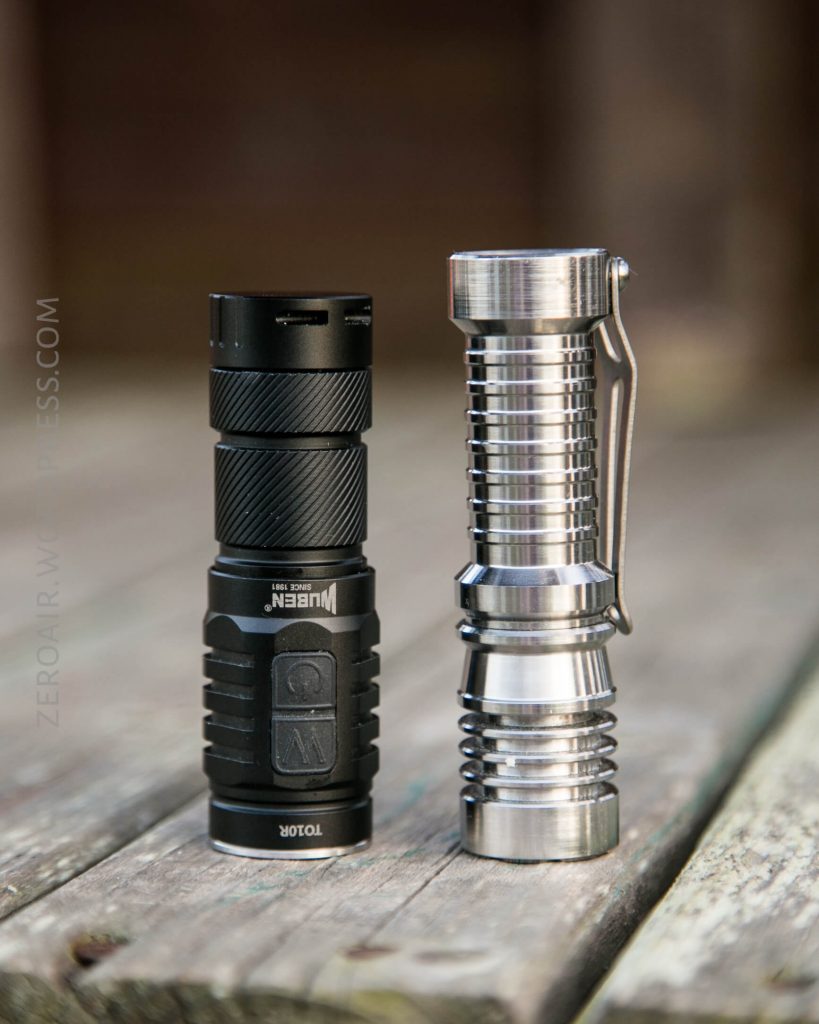 Two flashlights stand upright on a wooden surface. The flashlight on the left is black with rubberized sections, while the one on the right is silver with a metallic, grooved design and a side clip.