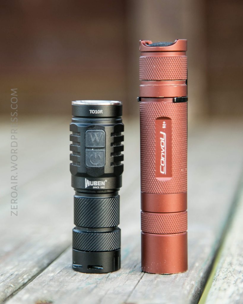 Two compact flashlights stand upright on a wooden surface. The left flashlight is black with WUBEN TO10R labeling, and the right one is red with Convoy branding. The background is blurred.