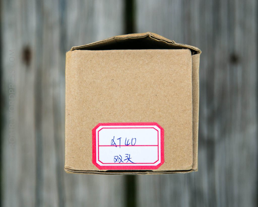 A brown cardboard box viewed from above, with a red and white label featuring handwritten text in blue ink. The background is a wooden surface with vertical lines.
