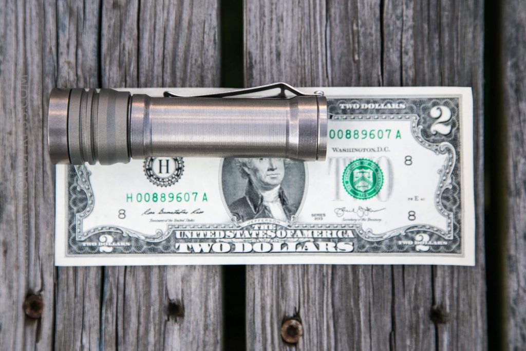 A metallic flashlight is placed horizontally across a two-dollar bill on a wooden surface with visible gaps between the planks.
