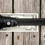 A black WOWTAC A4 flashlight is placed on top of a two-dollar bill on a weathered wooden surface, showing the size comparison between the flashlight and the bill.