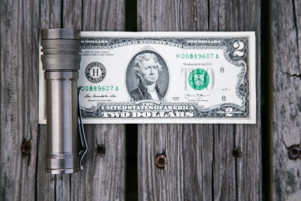 A flashlight is placed next to a U.S. two-dollar bill on a wooden surface with visible cracks and grain lines. The flashlight and bill are arranged parallel to each other.