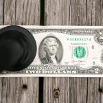 A two-dollar bill lies flat on a wooden surface with a black circular object, possibly a stamp or magnifier, partially covering the left side of the bill.