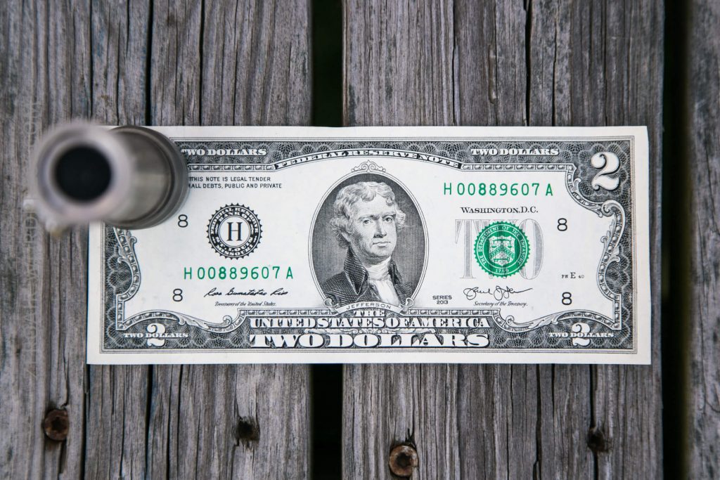 A U.S. two dollar bill featuring Thomas Jefferson is placed on a wooden surface with a metallic cylindrical object partially visible on the left side of the bill.