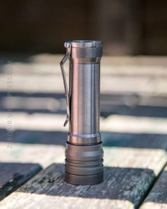 A metal flashlight with a pocket clip stands upright on a wooden surface with sunlight and shadows in the background.