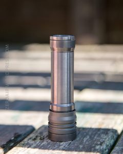 A metallic flashlight stands upright on a weathered wooden surface outdoors, with a blurred background and sunlight casting soft shadows.