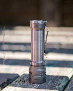 A metallic flashlight with a clip is standing upright on a wooden surface outdoors. The background is blurred and out of focus.