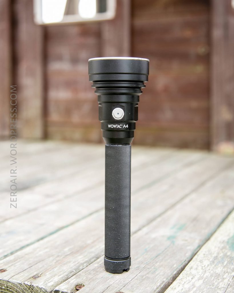 A black WOWTAC A4 flashlight is standing upright on a weathered wooden surface with a blurred wooden structure in the background.