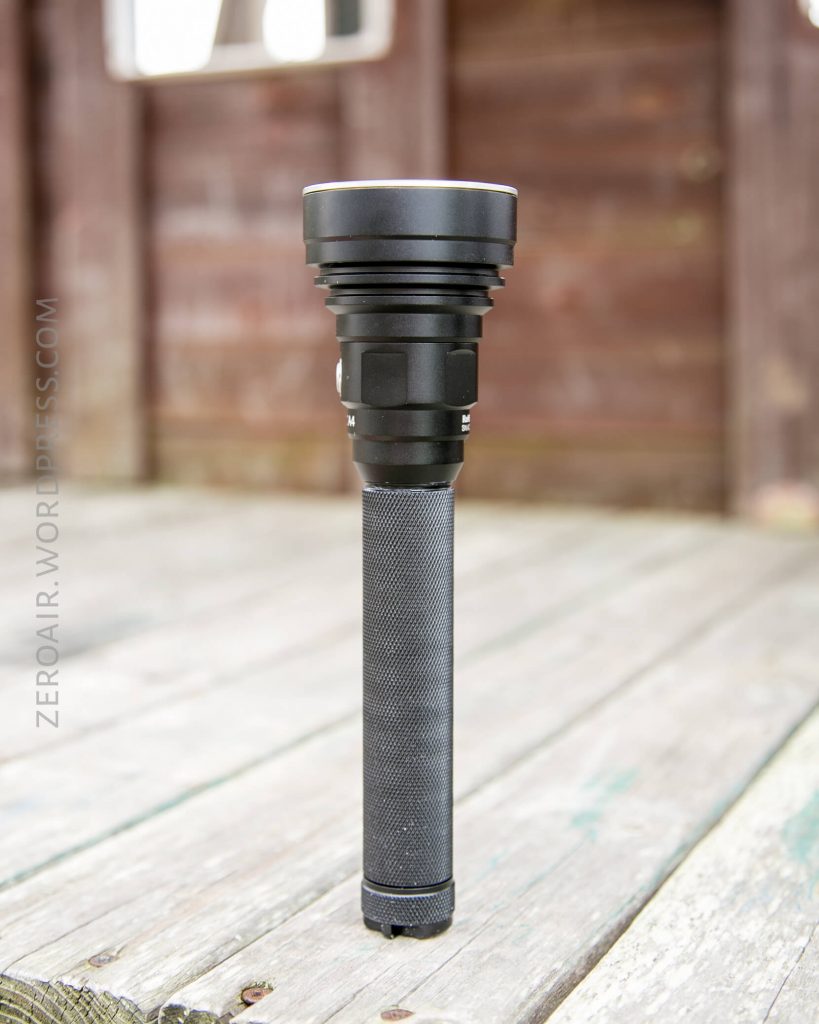 A black, textured flashlight stands upright on a weathered wooden surface with a blurred wooden structure in the background. The flashlight is centered in the image.