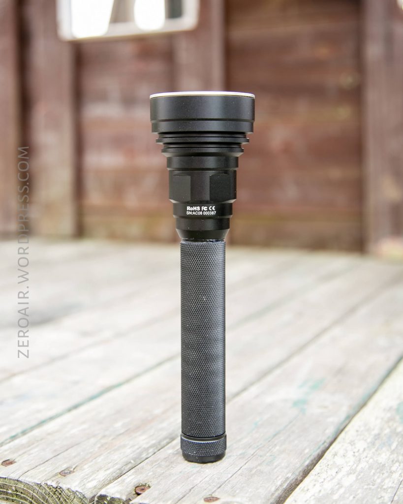 A large, black flashlight with a textured handle stands upright on a wooden surface outdoors, with an out-of-focus wooden wall in the background. The text ZEROAIR.WORDPRESS.COM is visible on the left side.