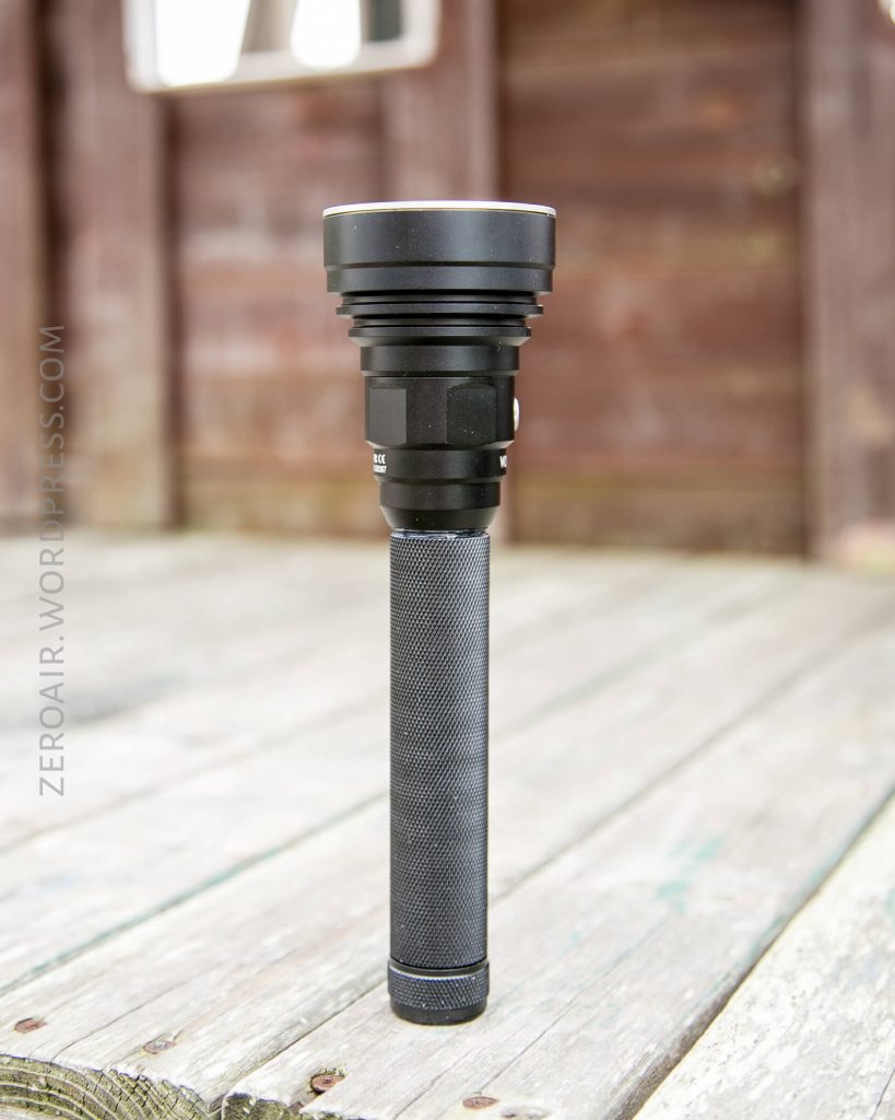 A large black flashlight with a textured handle stands upright on a wooden surface, with a blurred wooden structure in the background. The words “ZEROAIR.WORDPRESS.COM” are faintly visible along the left side.