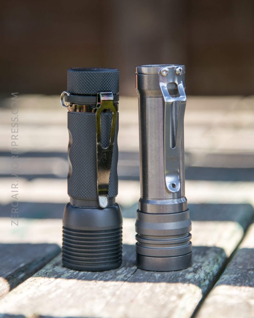 Two compact flashlights with metal clips stand upright side by side on a wooden surface. The left flashlight has a textured grip, while the right one has a smooth metallic finish.