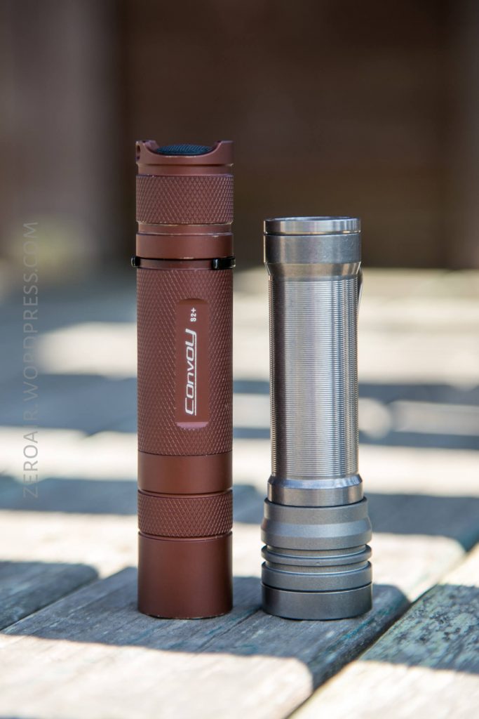 Two cylindrical flashlights stand upright on a wooden surface in sunlight. The left flashlight is brown with a Convoy label, and the right flashlight is metallic silver with a plain design.