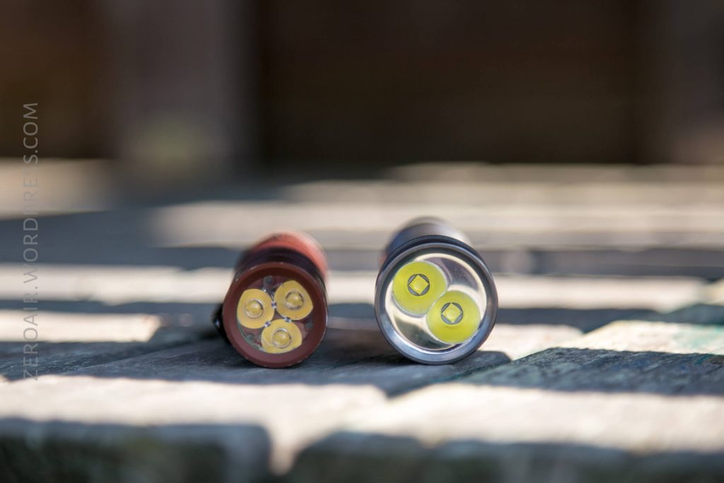 Two flashlights are lying side by side on a wooden surface, facing the camera. Each flashlight has three LED bulbs visible at the front. The background is out of focus.