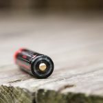 A single cylindrical battery with a black and red label is lying on its side on a weathered wooden surface, with the positive terminal facing the camera.