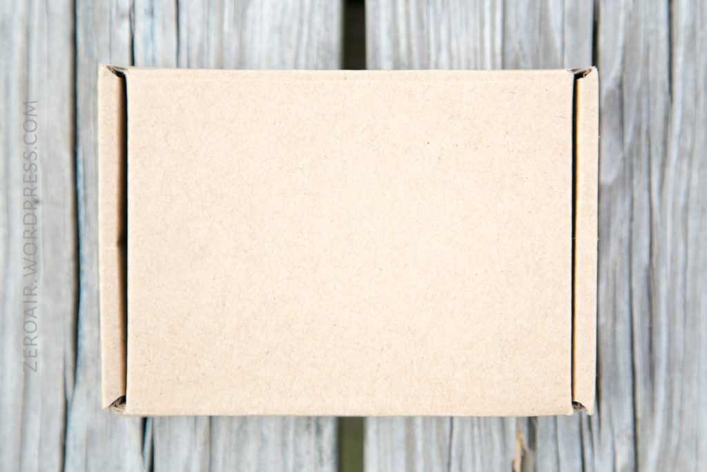 A plain, closed cardboard box sits on a weathered wooden surface. The box is centered in the image, and there is a faint website watermark on the left side.
