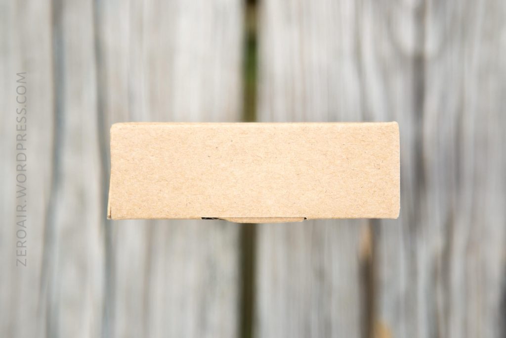 A plain, rectangular cardboard box viewed from the side against a blurred wooden background. The box appears closed and unmarked except for a faint watermark reading zeroair.wordpress.com on the left side.