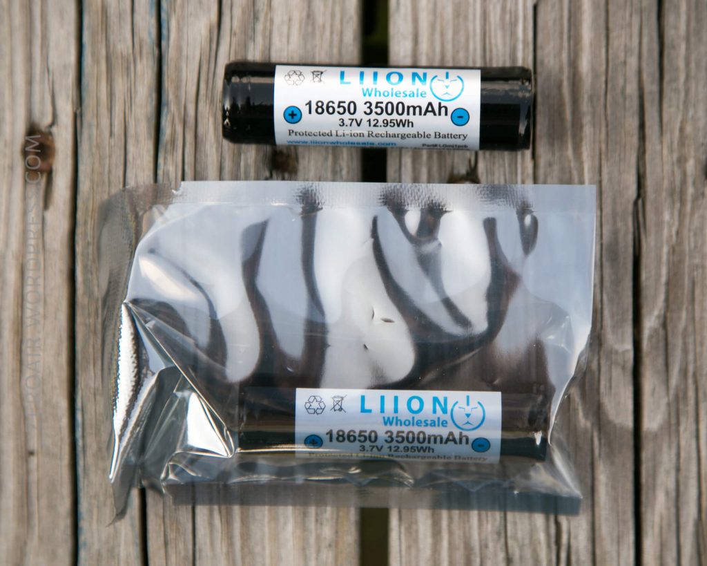 Two black 18650 3500mAh Li-ion rechargeable batteries are shown; one is unwrapped at the top, and the other is inside a clear plastic bag with a label, all placed on a wooden surface.
