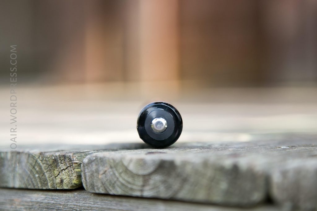 A close-up of a black cylindrical object, possibly a battery, lying horizontally on weathered wooden planks with a blurred background. The image is focused on the round end of the object.