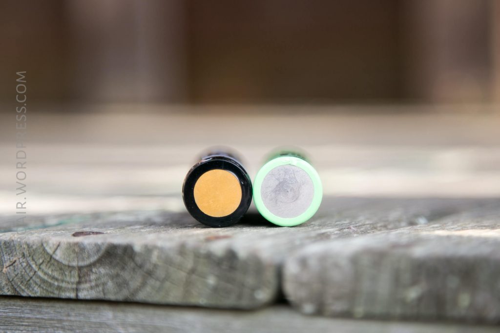 Two cylindrical batteries are lying on their sides on a wooden surface, with the positive ends facing the camera. The battery on the left has a yellow tip; the one on the right has a green tip.