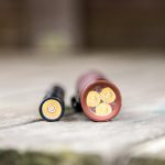 A close-up of two batteries lying horizontally on a wooden surface, viewed from the contact end. The left battery is smaller and black, while the right battery is larger and red with four visible contact points.