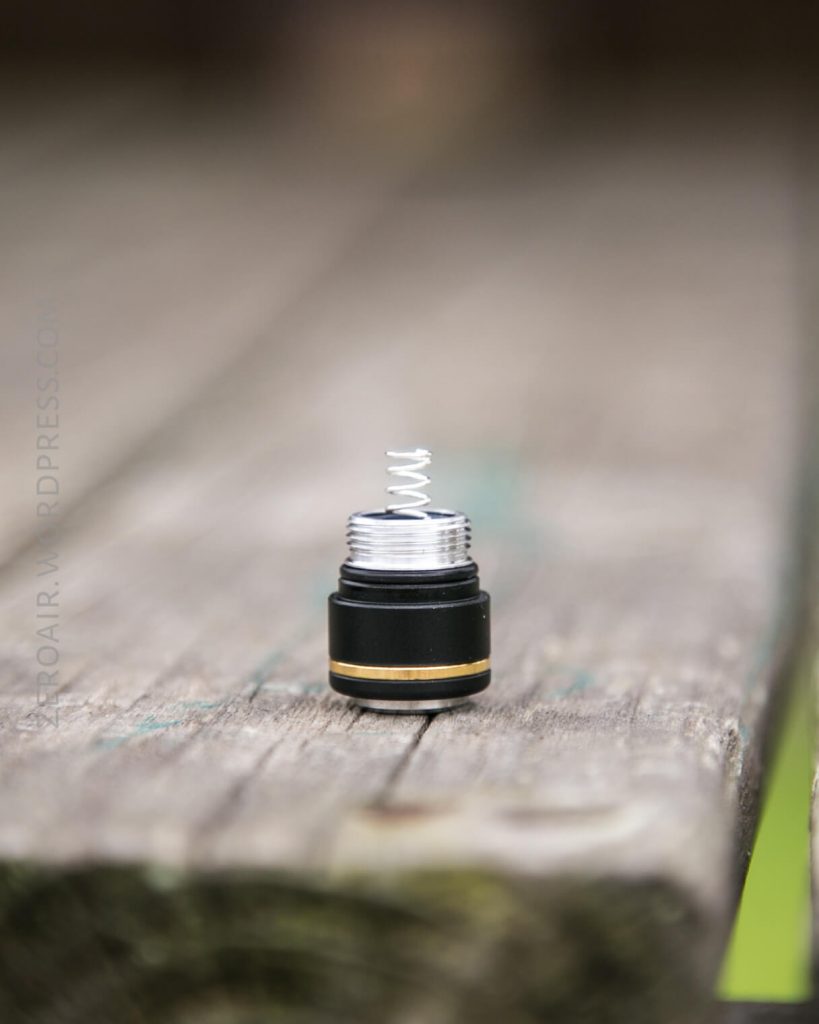 A close-up of a small black and silver cylindrical object with a visible metal spring on top, placed upright on a weathered wooden surface. The background is blurred.