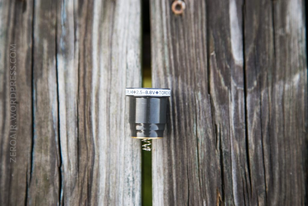 A small cylindrical electronic component with a spring at the bottom is mounted vertically in a gap between weathered wooden planks. The label on the component includes text and numbers.