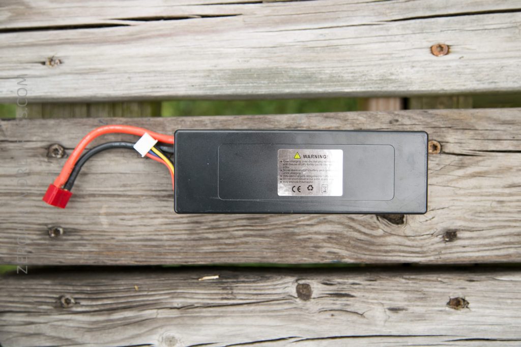 A rectangular black battery pack with a warning label, several wires, and a red connector sits on a weathered wooden surface.