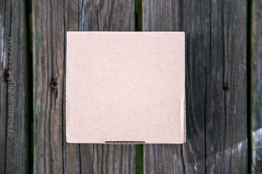 A plain, closed cardboard box sits on a weathered wooden surface, viewed from above. The box is centered in the image, and the wood shows visible grain and dark lines.
