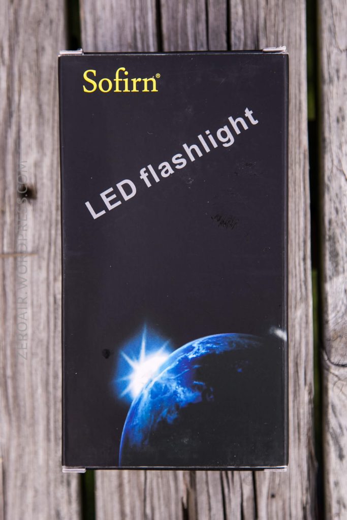A black Sofirn LED flashlight box is shown on a wooden surface. The box features an image of Earth with a bright light on the horizon and the words Sofirn LED flashlight printed on it.