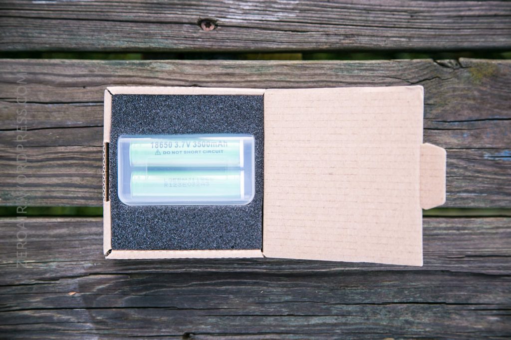 A cardboard box with foam padding contains two cylindrical green batteries labeled 18650 3.7V 3500mAh placed side by side, resting on a wooden surface. The box lid is partially open.